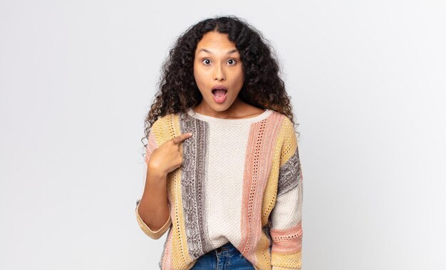 Pretty hispanic woman looking shocked and surprised with mouth wide open, pointing to self