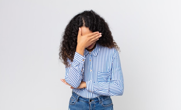 Foto bella donna ispanica che sembra stressata, imbarazzata o turbata, con mal di testa, che copre il viso con la mano