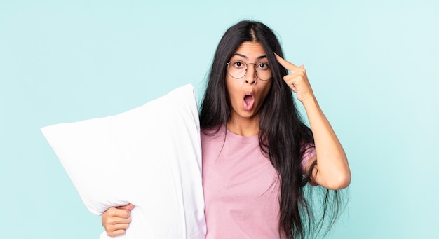 Pretty hispanic woman looking surprised, realizing a new thought, idea or concept and wearing pajamas with a pillow