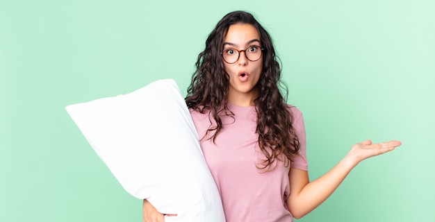 Pretty hispanic woman looking surprised and shocked, with jaw dropped holding an object and wearing pajamas with a pillow