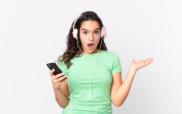Pretty hispanic woman looking surprised and shocked, with jaw dropped holding an object with headphones and a smartphone