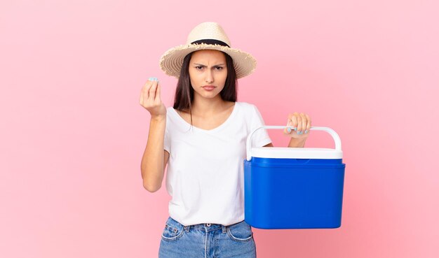 Pretty hispanic woman making capice or money gesture, telling you to pay and holding a portable refrigerator