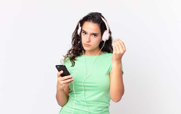 Pretty hispanic woman making capice or money gesture, telling you to pay with headphones and a smartphone