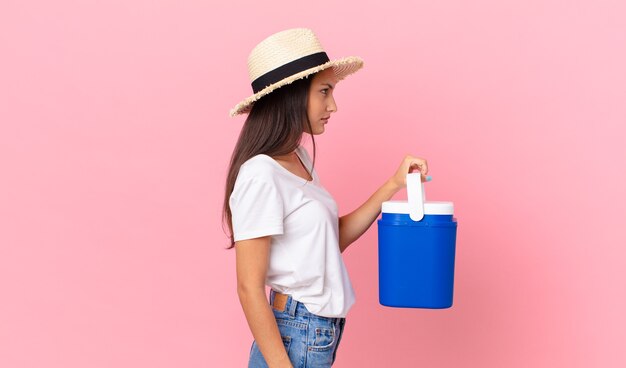 Pretty hispanic woman on profile view thinking, imagining or daydreaming and holding a portable refrigerator