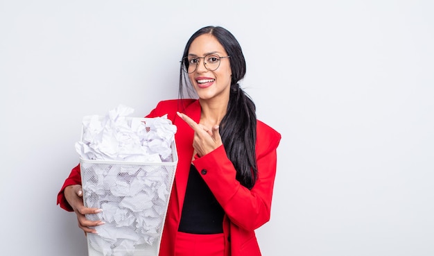 Pretty hispanic woman smiling cheerfully, feeling happy and pointing to the side paper balls trash concept