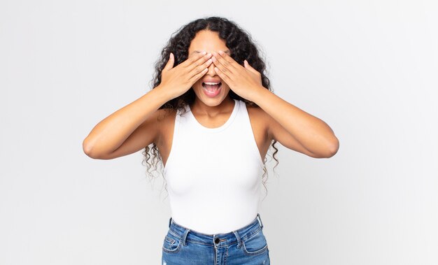 Pretty hispanic woman smiling and feeling happy, covering eyes with both hands and waiting for unbelievable surprise