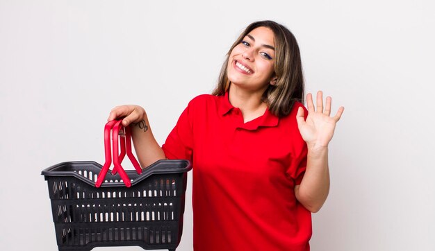 Pretty hispanic woman smiling happily waving hand welcoming and greeting you empty shopping basket concept