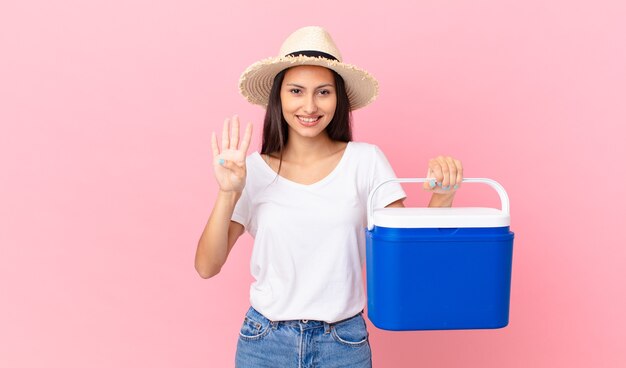 Pretty hispanic woman smiling and looking friendly, showing number four and holding a portable refrigerator