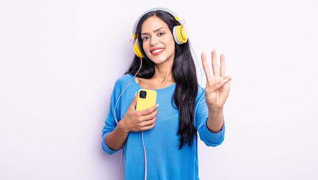 Pretty hispanic woman smiling and looking friendly, showing number three. smartphone and headphones concept