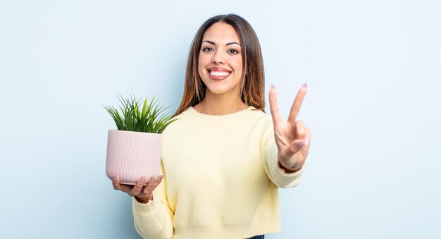 Pretty hispanic woman smiling and looking friendly, showing number two. houseplant concept