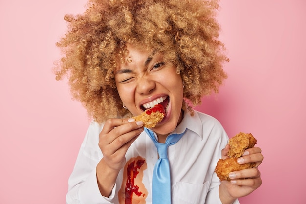 Pretty hungry female model eats fried nuggets with ketchup\
enjoys high calorie fast food dressed in white formal shirt and tie\
poses against pink background binge eating unhealthy nutrition