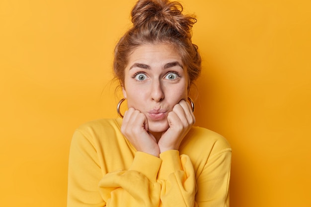 Pretty impressed woman keeps lips folded stares with widely opened eyes keeps hands under chin reacts on something exciting dressed casually isolated over vivid yellow background. Face expressions