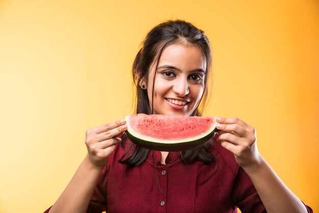 Pretty Indian or asian girl holding a slice of watermelon or Tarbuja in Hindi, standing isolated over white or yellow background.