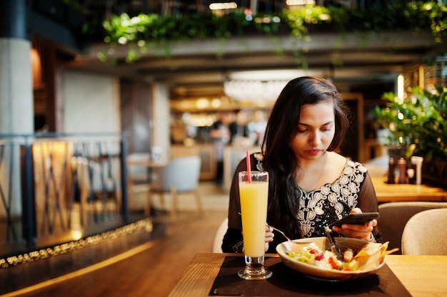 Красивая индийская девушка в черном платье сари позирует в ресторане, сидя за столом с соком и салатом, глядя на мобильный телефон