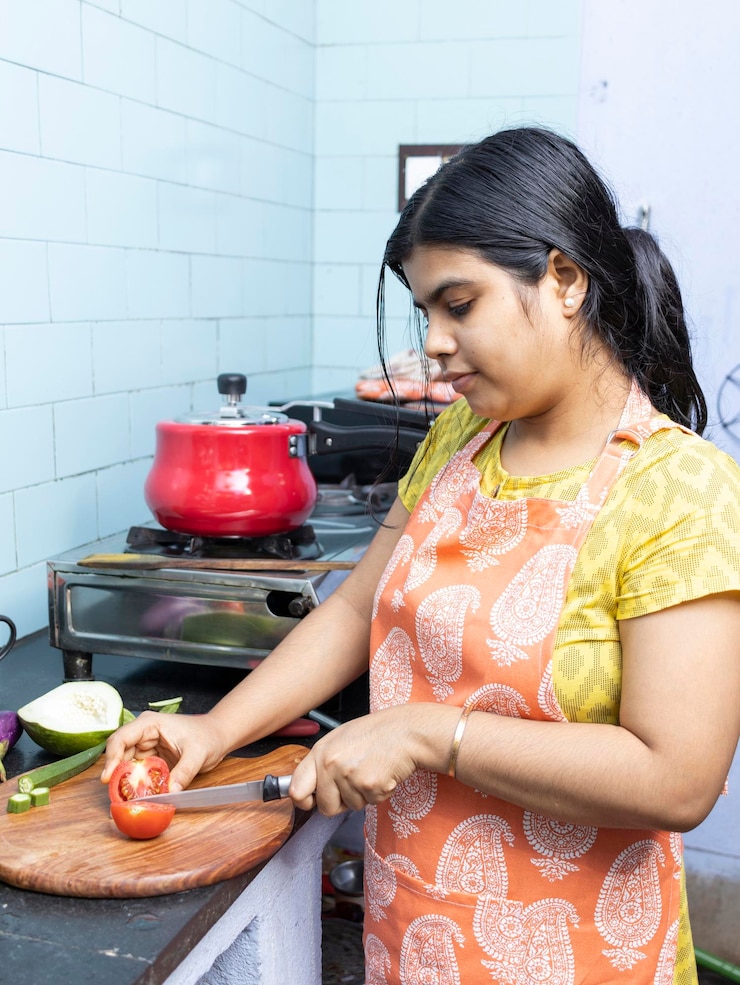 फोटो a pretty indian young woman wearing apron cooking and cutting vegetables in domestic kitchen