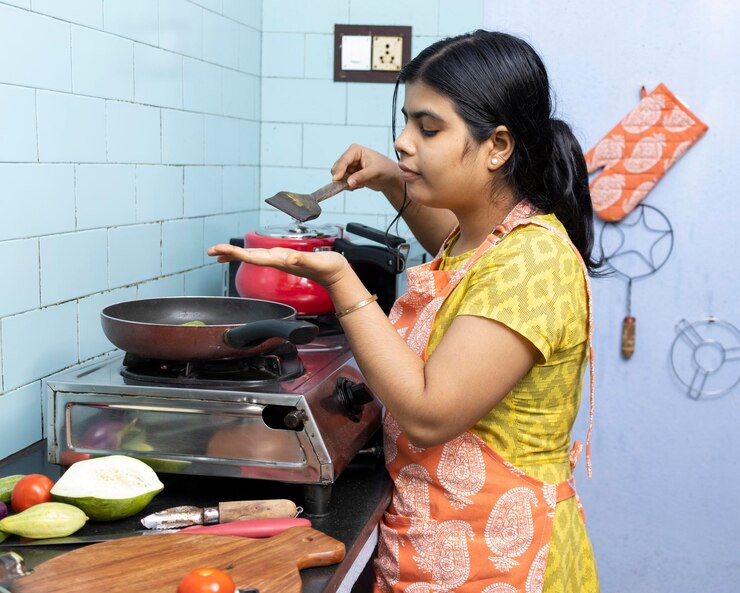 फोटो a pretty indian young woman wearing apron cooking and tasting food in domestic kitchen on gas stove