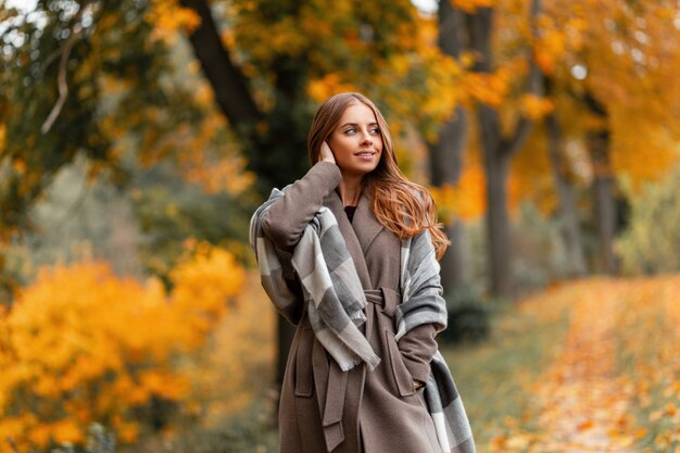 Pretty joyful young woman model in a stylish coat with a knitted vintage scarf posing in a forest on a background of trees with orange foliage. Attractive happy girl relaxes outdoors in the park.