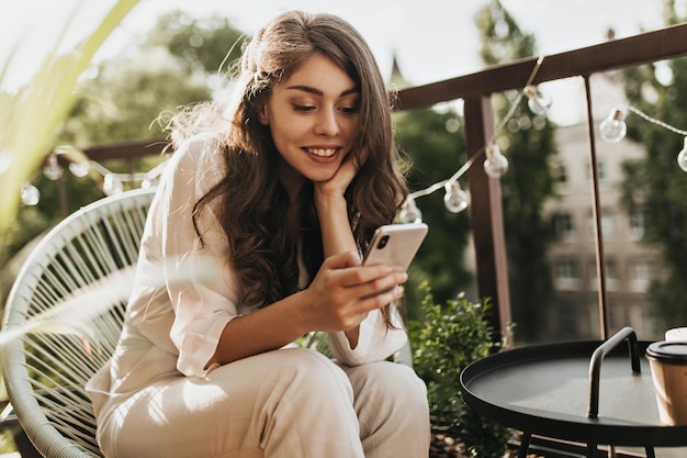 Pretty lady in beige outfit resting on terrace and holding phone Charming brunette woman in white blouse and pants chilling on balcony