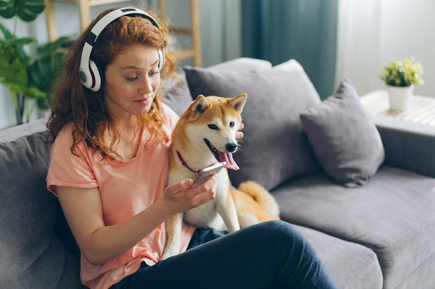 Bella signora che si gode la musica in cuffia utilizzando lo smartphone che accarezza il cane a casa