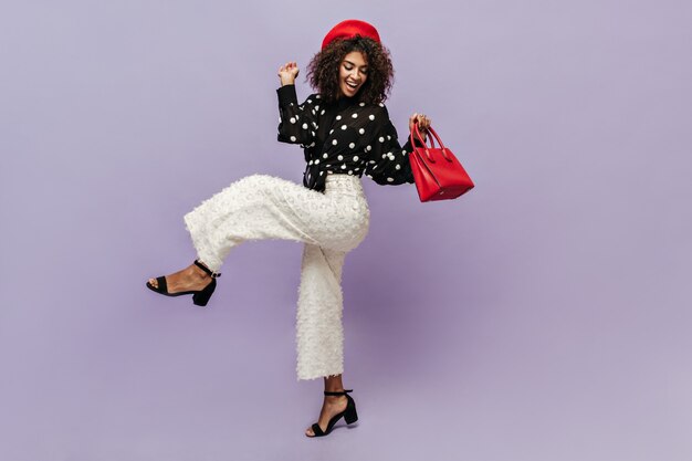 Pretty lady in good mood with dark curly hair in black modern blouse and wide white pants smiling and posing with red handbag