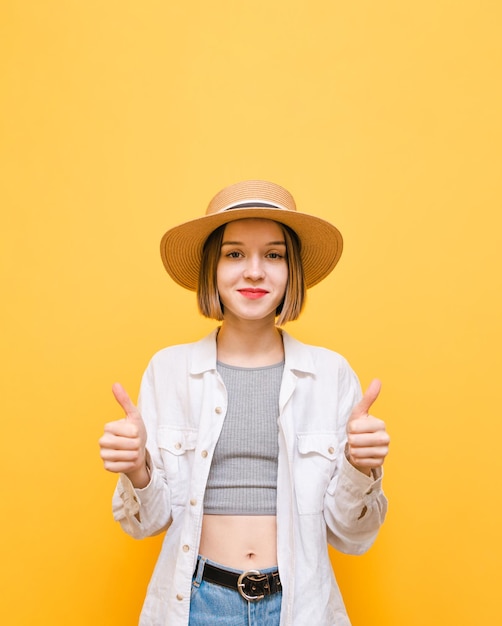 Pretty lady in hat and light clothing looks into camera and shows thumbs up on yellow background