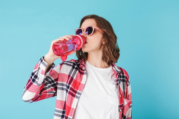 Pretty lady in sunglasses standing with pink sport bottle and drinking water over colorful background