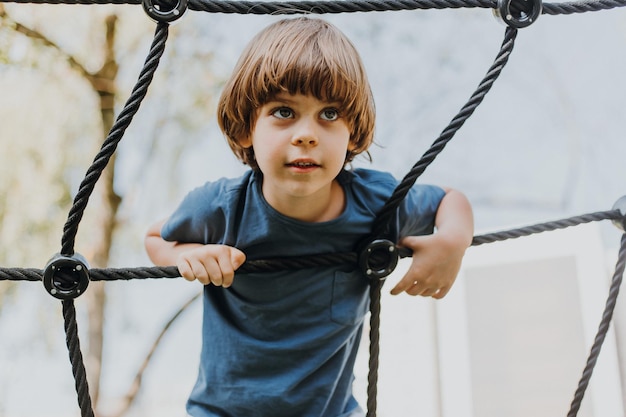 Pretty little brunette boy in a blue t-shirt is climbing a rope\
web. child is engaged in outdoor sports. healthy active kid.\
lifestyle. space for text. high quality photo