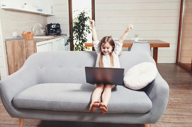 Photo pretty little brunette girl sitting on the sofa and playing games on the laptop winner gesture