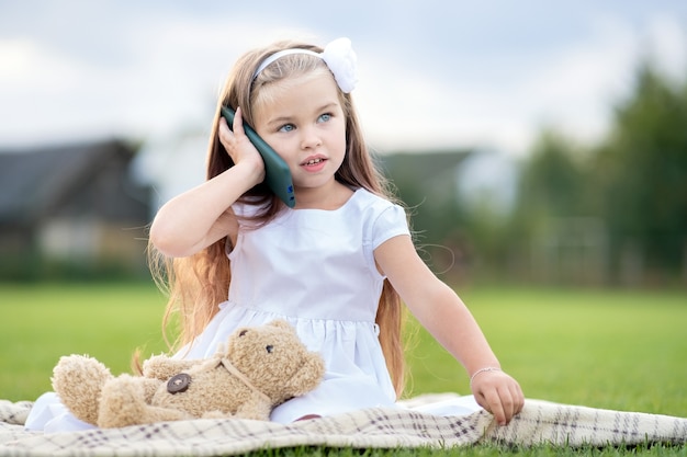 휴대 전화로 이야기하는 그녀의 테디 베어 장난감과 함께 여름 공원에 앉아있는 예쁜 어린 소녀