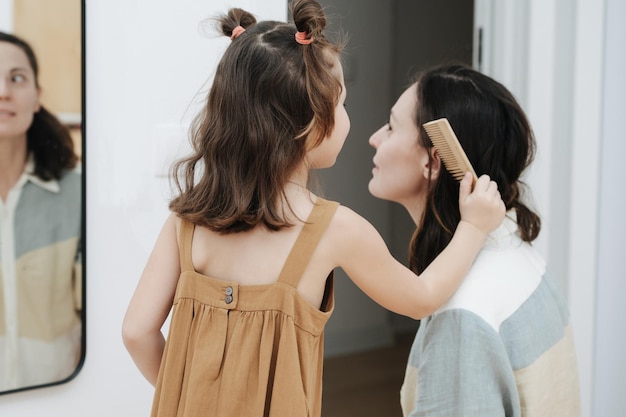 Pretty little girl brushing mother's hair in front of a mirror Her mom is surprised