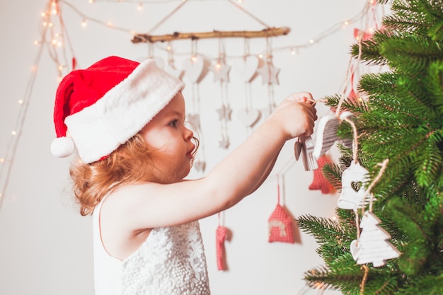 Pretty little girl in a cap of Santa Claus is decorating Christmas tree