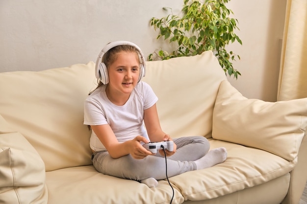 Pretty little girl in headphones are playing game console, looking on the screen and smiling while sitting on sofa at home