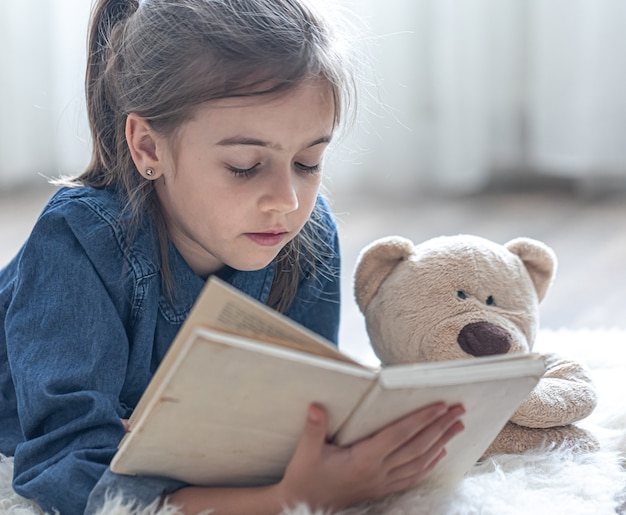 Graziosa bambina a casa, sdraiata sul pavimento con il suo giocattolo preferito e legge un libro.