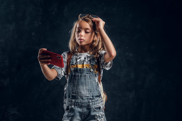 Pretty little girl is making selfie by mobile phone at dark photo studio.