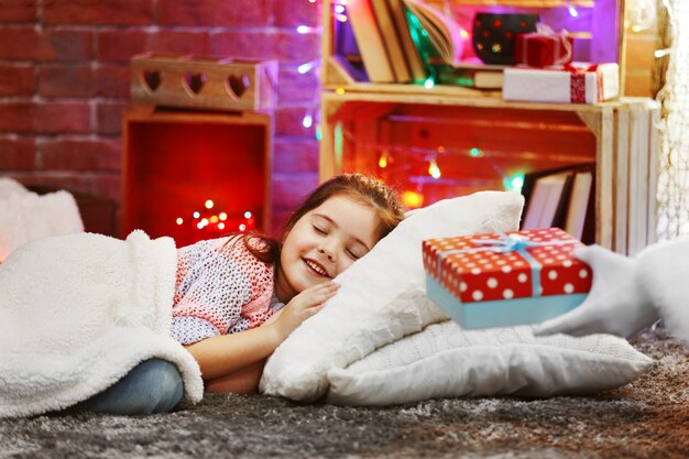 Foto graziosa bambina sdraiata sul cuscino sotto un morbido plaid in attesa del regalo di babbo natale nella stanza decorata di natale