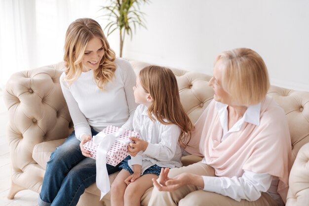 Pretty little girl passing a gift box tied up with a white ribbon to her mother while her grandmother looking at this process