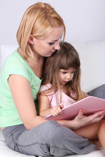 Pretty little girl reading book with mother on sofa on gray background