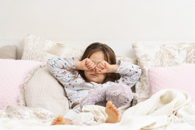 Pretty little girl in sleepwear lying on bed and rub eyes with fists Portrait of awaking child Kid wake up at morning