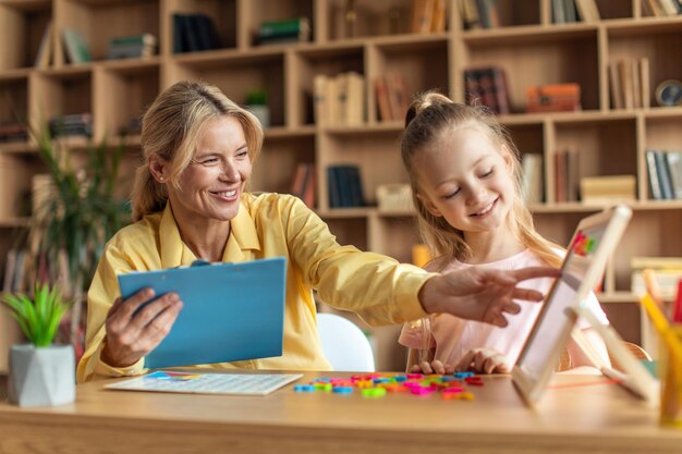 Pretty little girl studying to read with professional female tutor friendly woman teacher helping kid and taking notes