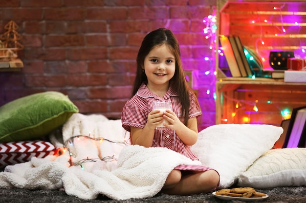 Pretty little girl with glass of milk and tasty cookies in Christmas decorated room