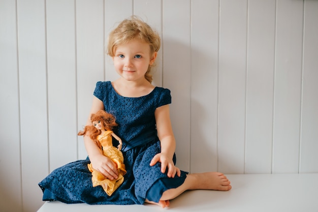 Pretty little girl with short fair hair in blue dress holds her lovely barbie toy, sits in bright baby room and smiles
