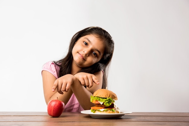 Pretty little Indian or asian girl unable to choose between fresh apple and burger, healthy eating concept