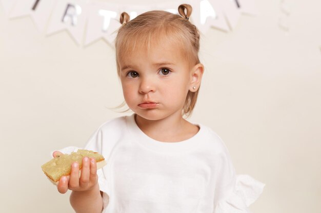 Pretty little infant girl with bun hair buns dressed white attire enjoys delicious cake for her birthday posing against white wall looking at camera Children's holiday Happy childhood