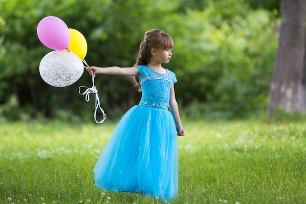 Pretty little long-haired girl in long blue dress holding colorful balloons 