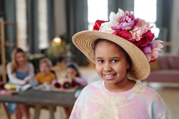 Pretty little mixed-race girl in hat decorated with large handmade flowers of white, pink and crimson colors standing against her friends by table