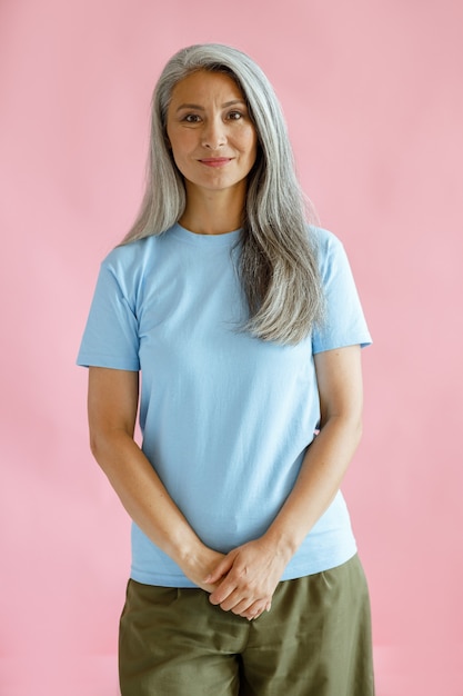 Pretty long haired middle aged Asian lady in blue t-shirt stands on pink background in studio. Mature beauty lifestyle