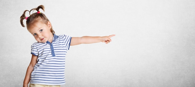 Photo pretty lovely beautiful small kid wears striped t shirt, has funny two pony tails, pointing with her little hand