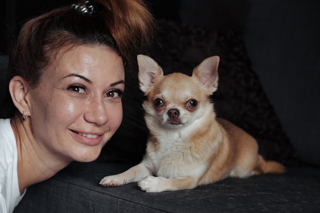 Donna abbastanza matura con il cane chihuahua sul divano scuro nel soggiorno di casa. femmina di mezza età e il suo cagnolino chihuahua. concetto di amore per animali domestici e amico di famiglia