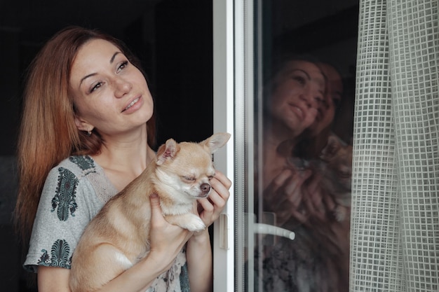 Pretty mature woman with Chihuahua dog in hands on home balcony. Middle aged female and her doggy Chihuahua. Concept pet love and family friend