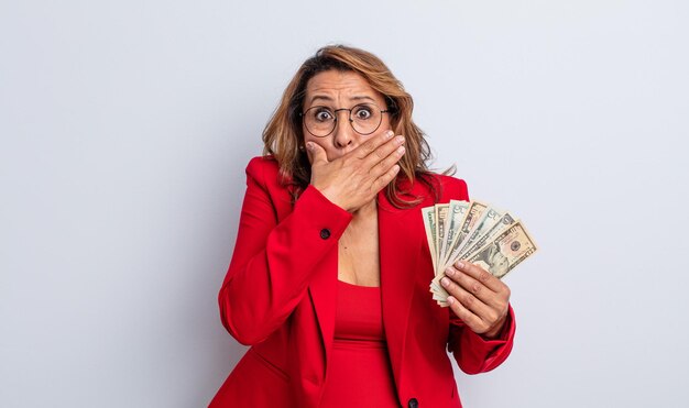 Pretty middle age woman covering mouth with hands with a shocked. dollar banknotes concept